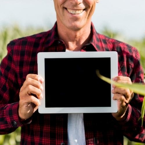 Imagem de um homem sorrindo vestindo camisa xadrez e blusa branca segurando um tablet com a tela voltada para você, ele está em uma plantação